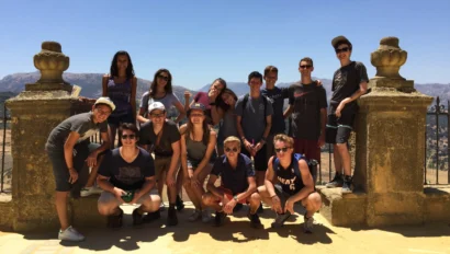 A group of thirteen people from the Spain summer travel program poses together outdoors on a sunny day, standing and kneeling in front of a stone railing with a scenic background.