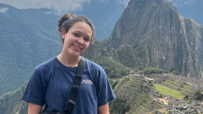 A young woman on a Peru student travel adventure stands in front of the ancient Incan city of Machu Picchu, with terraced ruins and a mountain peak visible in the background under cloudy skies.