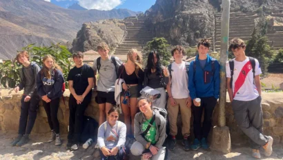 A group of young people pose in front of mountain ruins under a sunny sky, standing and sitting on a stone path.