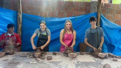Four people at a pottery workshop, each shaping clay on a table. They are under a blue tarp with red brick walls in the background.