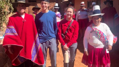 A group of people stands outdoors. Some wear traditional clothing, including red garments and hats, while others wear casual attire. They are on a sunny day near a rustic building.
