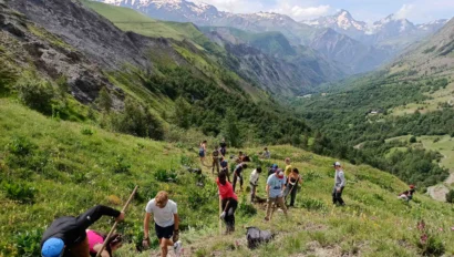 A group of people hike up a grassy mountain slope with hiking poles, surrounded by scenic views of a valley and distant snow-capped peaks under a cloudy sky.