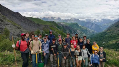 A group of people with tools poses on a grassy hill with mountains in the background.