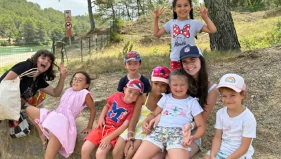 A group of children and one adult sit and pose for a photo outdoors, surrounded by trees and wearing casual summer clothes and hats during a fun French immersion program.
