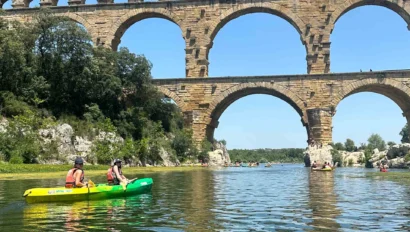 Two kayakers paddle on a river beneath an ancient stone arch bridge under a clear blue sky.