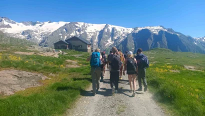 A group of hikers walks on a dirt trail toward a cabin, with snow-capped mountains in the background under a clear blue sky.