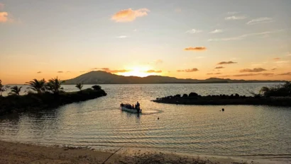 A small boat with people on calm water during a sunset, surrounded by palm trees and distant mountains.