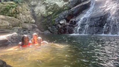 Three people swimming in a natural pool under a small waterfall surrounded by rocks and greenery.