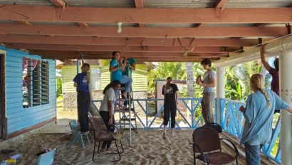 People renovating a covered patio by the sea, working on the roof and railings. Chairs and tools are scattered around.