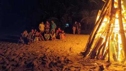 A group of people sitting on a sandy beach at night near a large bonfire.