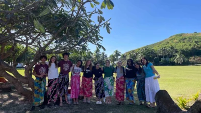 A group of people wearing colorful skirts stands together under a tree, with a grassy field and tropical landscape in the background.