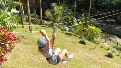 A person wearing a helmet and gloves rides a zipline over a grassy area surrounded by trees and plants in daylight, capturing the thrill of adventure on a Fiji student trip.
