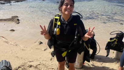 A person in a wetsuit and scuba gear stands on a sandy beach, smiling and making peace signs. Ocean waves are visible in the background.