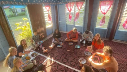 A group of people sit on a decorated floor in a room with red curtains, sharing a meal from plates placed in front of them.