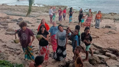 A group of people, including children and adults, stand on a rocky beach with the ocean and boats in the background. They appear to be gathered near a palm tree.
