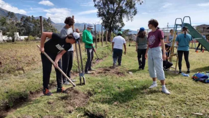 A group of people working outdoors, digging and preparing the ground in a grassy area with shovels and tools. Some are gathered around, while others are working individually. Trees and playground equipment are visible.