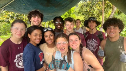 A group of smiling people wearing gloves and safety glasses stand under a tarp outdoors. Some have thumbs up. They're surrounded by greenery.