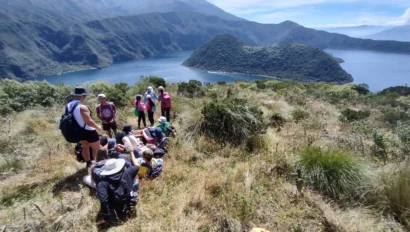 Group of hikers resting on a grassy hill overlooking a lake and mountainous landscape under a clear sky.