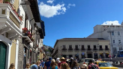 A group of people walk down a sunny street with historic buildings, taxis, and a blue sky.