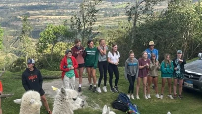 A group of people stands on a grassy hill with alpacas in the foreground and a scenic mountain landscape in the background.