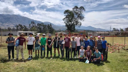 A group of people, including children and adults, stands outside on grass with a mountain backdrop. They appear to be in a park or field area.