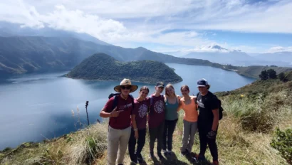 Group of people posing on a hilltop overlooking a lake and mountains under a partly cloudy sky.