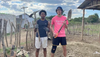 Two people stand outdoors holding tools, one with a pickaxe and the other with a shovel. A rural setting with a simple fence and clouds in the background.