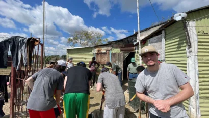 A group of people, some wearing hats, work together on a construction project outside a corrugated metal building under a blue sky with scattered clouds. A man stands in the foreground.