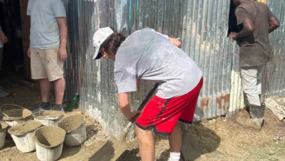 A person in a gray shirt and red shorts shovels cement near a corrugated metal wall. Two others stand nearby, one watching and the other working at a window. Buckets of cement are on the ground.