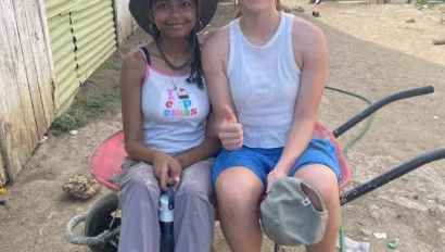 Two people sit side by side on a red wheelbarrow in a rural area of the Dominican Republic, with corrugated metal structures and banana trees behind them—one gives a thumbs-up during their Pre Med travel program experience.
