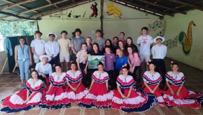 A group of people pose indoors, with some wearing traditional red, white, and blue dresses. The backdrop features musical and theatrical artwork.
