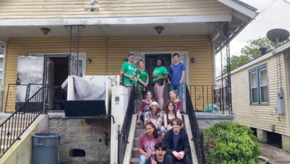 A group of people sit and stand on the steps of a weathered house, some wearing green shirts. A washing machine and a trash can are nearby.