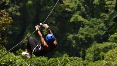 Person zip-lining through a lush green forest, wearing a blue helmet, gloves, and casual clothing.