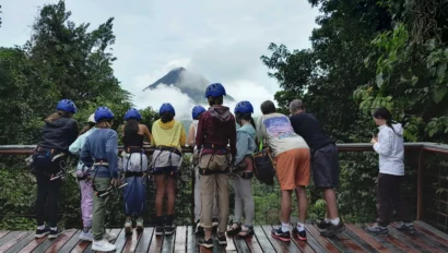 A group of people in helmets and harnesses stand on a wooden platform, looking at a cloud-covered mountain in a lush forest setting.