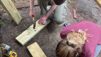 Two people crouch on the ground, assembling wooden beams with drills and a hammer. Construction tools are scattered around them.