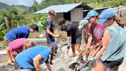 A group of people are using shovels to mix and move sand or gravel at a construction site in a rural, hilly area. They appear to be working together on a project outside.