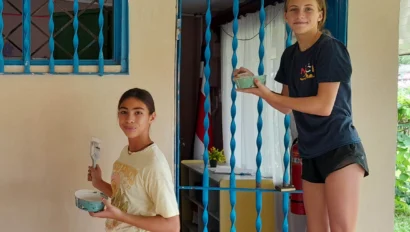 Two young people on a Costa Rica student travel experience paint the outside wall and metal bars of a building. One stands on a chair with a paint tray, while the other adds color to the wall.
