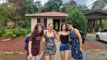 Four people stand smiling in front of a house with a garden. One wears a patterned dress, others wear casual outfits, and two have backpacks. There’s a white car to the side.