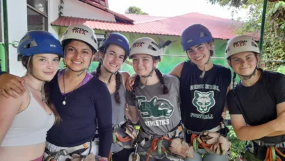 Six people wearing helmets and harnesses stand together under a canopy, smiling at the camera. They appear ready for an outdoor adventure activity.