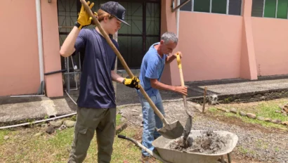 Two people are mixing concrete with shovels and a wheelbarrow in an outdoor setting near a building.