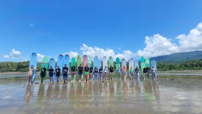A group of people with surfboards stands in a line on a sandy beach, with a clear blue sky and mountains in the background.
