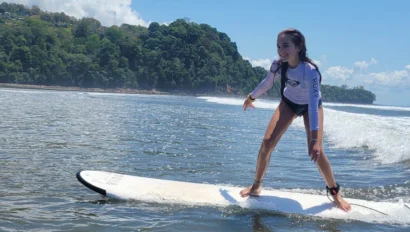 A person is surfing on a white surfboard near the shore, with green trees and a blue sky in the background—a perfect scene for Costa Rica student travel adventures.