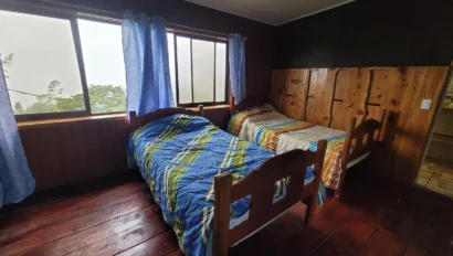 A small wooden bedroom with two single beds, each covered with different blankets. A large window with blue curtains overlooks greenery outside.