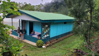 A small blue house with a green roof surrounded by lush greenery. Two people are on the porch, and various potted plants are visible.