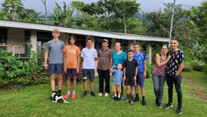 A group of eleven people, including adults and children, stand outside on grass in front of a single-story building during a language immersion trip, with a black and white dog lying in front.