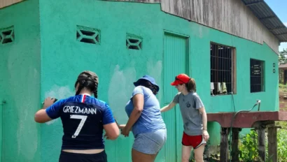 Three people repaint a green exterior wall of a building on stilts under a clear sky.