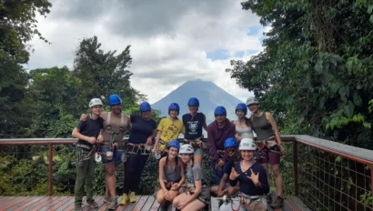 A group of people wearing helmets and harnesses pose on a wooden platform with a mountain and forest in the background.