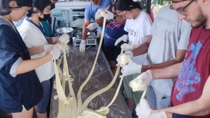 A group of people wearing gloves work together to stretch long strands of dough on a large table in a bright indoor space as part of an environmental school travel program.