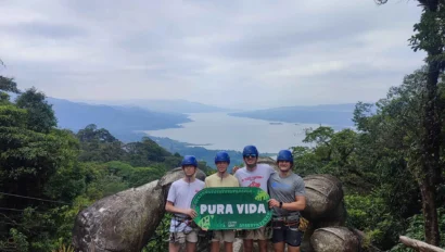 Four people wearing helmets stand on a large hand-shaped stone structure, holding a "Pura Vida" sign—a perfect moment from their environmental school travel program, surrounded by lush greenery and a scenic lake in the background.