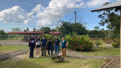 A group of six people stands outdoors on grass near a wheelbarrow, participating in an environmental school travel program, with greenery, a fence, and clouds in the background.
